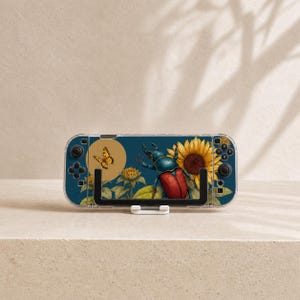 Op de afbeelding: Een draagbare spelcomputer met een donkerblauwe skin met een botanische illustratie van een grote kever, een gele vlinder en felgele zonnebloemen. Het apparaat heeft zwarte knoppen, joysticks en een doorzichtige beschermhoes.