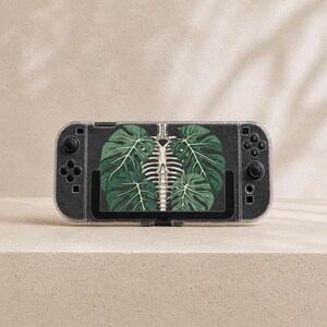 May include: A handheld gaming console featuring a black skin with a central graphic of a human rib cage intertwined with large green monstera leaves. The device has black joysticks, directional buttons, and action buttons on both sides.