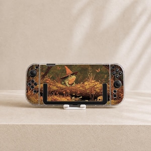 May include: A handheld gaming console featuring a skin with a green frog wearing a pointed orange hat, perched on a fallen log in a forest setting with mushrooms and a skull, framed by black controller buttons and joysticks on both sides.