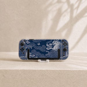 May include: A handheld gaming console featuring a dark blue protective skin with a white line-art design of a dragon, swirling clouds, and butterflies. The device has a clear outer shell, black buttons, and joysticks, and is mounted on a stand.