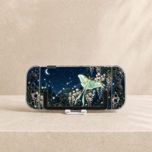 May include: A handheld gaming console featuring a clear protective case with a printed design of a pale green luna moth, white wisteria flowers, and a crescent moon against a dark blue night sky filled with glowing fireflies and forest trees.