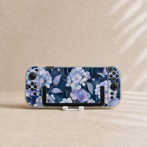 May include: A handheld gaming console featuring a dark blue skin with a pattern of light blue and purple hydrangea flowers and small white butterflies. The device has black joysticks, directional buttons, and action buttons on both sides.