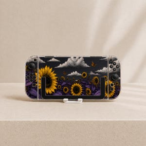 May include: A handheld gaming console featuring a protective skin with a dark background, yellow sunflowers, purple foliage, and white clouds. The device includes black joysticks, directional buttons, and action buttons on the sides of the screen.