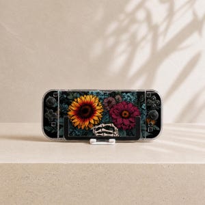 May include: A handheld gaming console featuring a protective skin with a dark background, a large yellow sunflower, a deep pink zinnia, and a skeletal hand graphic. The device has black buttons and joysticks on both sides of the screen.
