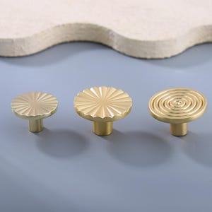 May include: Three gold-colored cabinet knobs with different designs. The knobs have a round shape with a stem. One knob has a sunburst pattern, another has a scalloped edge, and the third has a spiral design. The knobs are on a light blue surface.