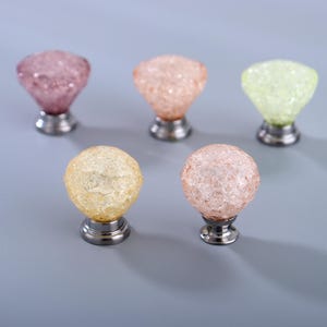 Könnte beinhalten: Fünf dekorative Schrankknöpfe in den Farben Rosa, Pfirsich, Gelb und Grün. Jeder Knopf hat ein facettiertes, kristallartiges Aussehen und einen silberfarbenen Sockel. Die Knöpfe sind auf einer hellgrauen Oberfläche angeordnet.
