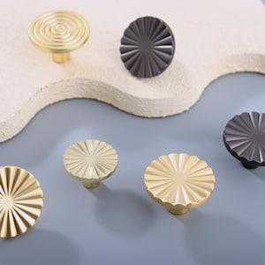 May include: A collection of decorative cabinet knobs in various designs and finishes. The knobs include gold and dark gray options, with radial and spiral patterns. These hardware pieces are designed for furniture and home decor.