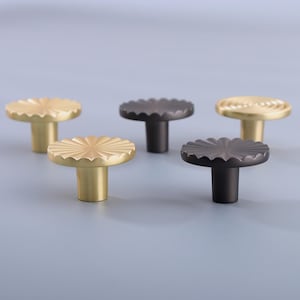 以下が含まれることがあります： 花のようなデザインの装飾キャビネットノブ5個。ノブは、金と濃いグレーを含むさまざまな金属仕上げです。ノブは、丸くスカラップされた上部と円筒形のステムを備えています。