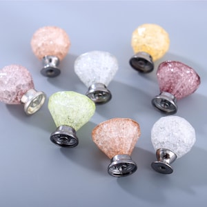 May include: A collection of eight decorative cabinet knobs in various colors, including pink, yellow, green, and clear. Each knob features a faceted, crystal-like design and a silver-toned base. The knobs are arranged on a gray surface.