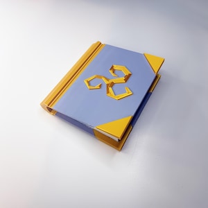 May include: A closed book with a light blue cover and gold accents. The cover features a gold emblem. The edges and corners of the book are also gold. The book is sitting on a white surface.