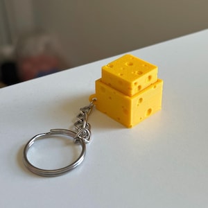 Puede incluir: Un llavero amarillo con forma de queso con un llavero plateado. El queso es un diseño de dos niveles con agujeros, que se asemeja al queso suizo. El llavero está sobre una superficie blanca.