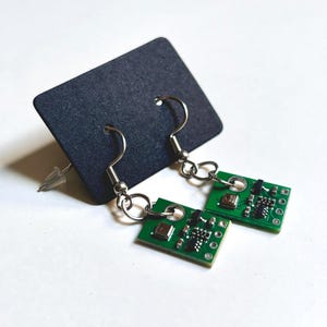 May include: A pair of unique earrings featuring small green circuit boards with silver-tone ear wires. The earrings are displayed on a black earring card against a white background. The circuit boards have various electronic components.
