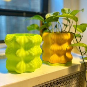 May include: Two decorative, wavy-edged planters. One is bright lime green, the other is yellow. Both have a textured, ribbed design. Green leafy plants are growing out of the yellow planter. The planters are sitting on a white surface.