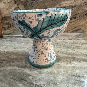 May include: A ceramic pedestal bowl with a white base and a speckled blue pattern. The bowl features a teal leaf design and a teal rim. This decorative bowl is suitable for serving or display.