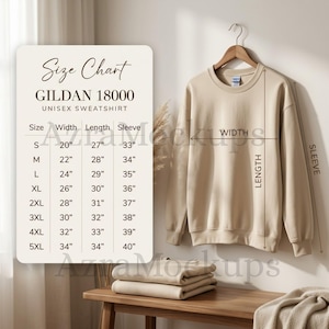 Puede incluir: Una sudadera unisex Gildan 18000 beige cuelga de una percha de madera. Una tabla de tallas muestra las medidas en cm para las tallas S a 5XL. Sudaderas dobladas y una manta descansan sobre una mesa de madera.