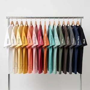 Puede incluir: Una colección de camisetas en varios colores, incluyendo blanco, marfil, mantequilla, mostaza, melón, flor, carmesí, naranja quemado, arrecife de la isla, espuma de mar, musgo, espresso, pimienta, azul marino y negro. Cada camiseta está en una percha de madera.