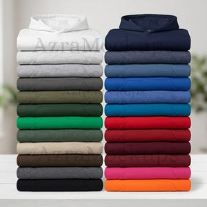 Könnte beinhalten: Gestapelte Kapuzenpullover in verschiedenen Farben, darunter Weiß, Grau, Grün und Schwarz. Die Kapuzenpullover haben eine Vordertasche und eine Kapuze. Das Bild zeigt eine Auswahl an Bekleidungsmöglichkeiten.