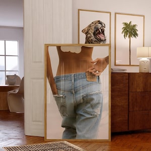 May include: A framed print showing a person in light blue denim jeans with distressed details. The image is reflected in a mirror, with leopard print and palm tree art in the background. The frame is made of light wood.