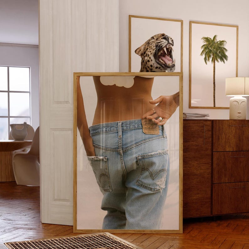 May include: A framed print showing a person in light blue denim jeans with distressed details. The image is reflected in a mirror, with leopard print and palm tree art in the background. The frame is made of light wood.