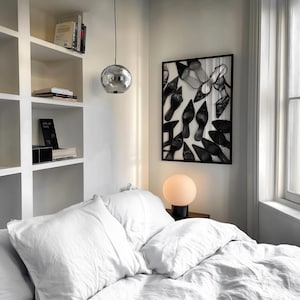May include: A bedroom scene featuring a white bed with pillows and a framed artwork of high heels. A round, silver pendant light hangs from the ceiling. A small, round lamp sits on a nightstand. Bookshelves are visible on the left.