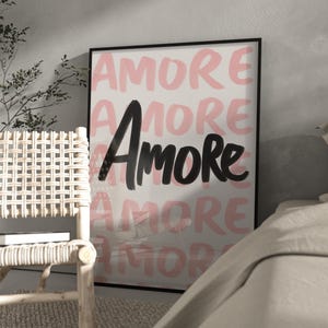 May include: A framed art print with the word "Amore" in a bold black font, layered over a repeating pattern of the same word in a soft pink hue. The artwork is set against a white background.