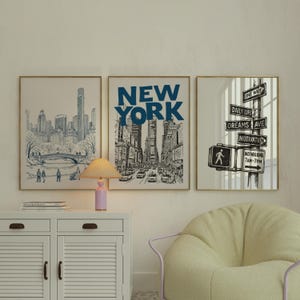 Puede incluir: Tres impresiones artísticas enmarcadas con marcos dorados. La impresión de la izquierda representa un boceto azul y blanco de un horizonte urbano con un puente. La impresión central dice "NEW YORK" en letras azules grandes. La impresión de la derecha muestra señales de tráfico.
