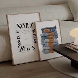 May include: Two framed prints. One features a clock face design with Roman numerals and the text "GIRL CLUB". The other print displays a stack of blue denim jeans with brown leather labels. Both prints are in light-coloured wooden frames.