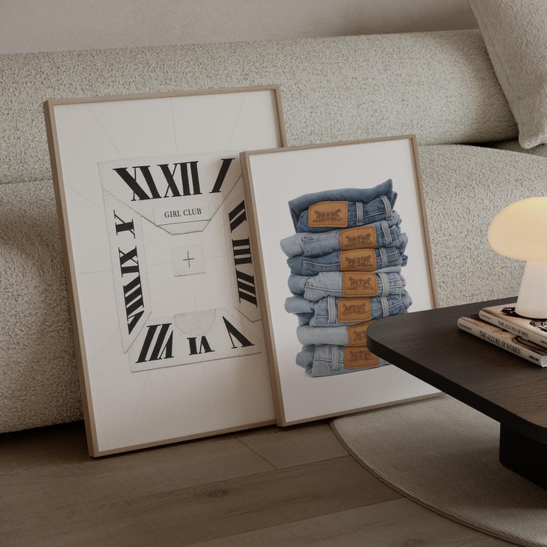 May include: Two framed prints. One features a clock face design with Roman numerals and the text "GIRL CLUB". The other print displays a stack of blue denim jeans with brown leather labels. Both prints are in light-coloured wooden frames.