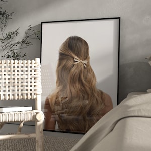 May include: A framed print showing a woman with long, wavy blonde hair styled with a small bow. The image is set against a white background, framed in black, and placed in a room with neutral tones. The print adds a touch of elegance to the decor.