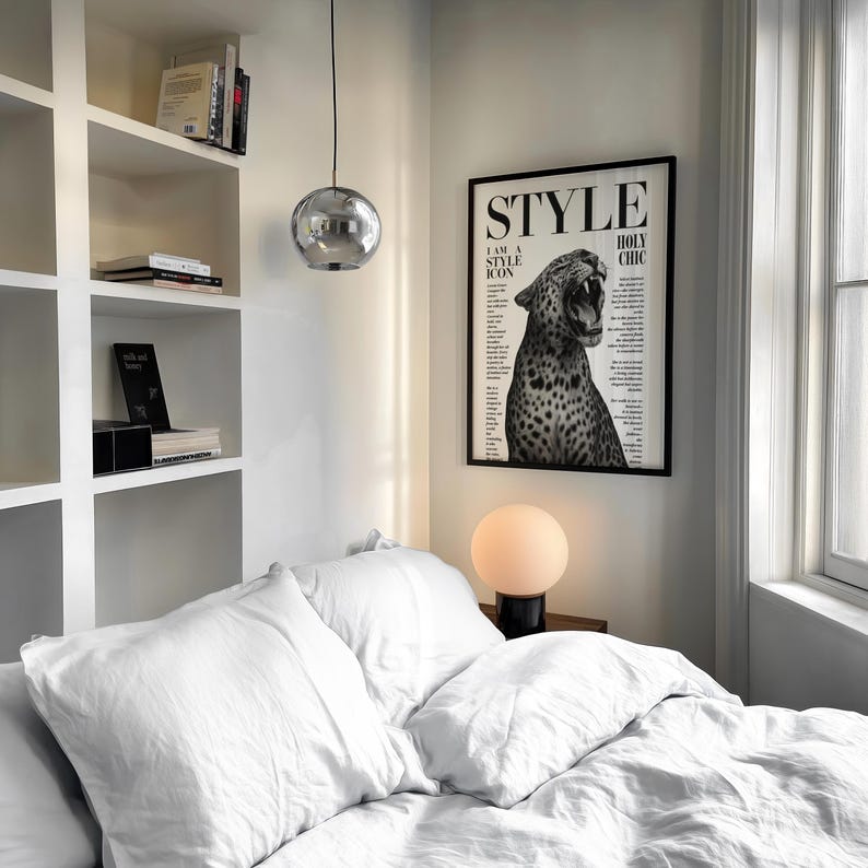 May include: A bedroom interior with a bed covered in white bedding, a framed black and white poster with the word "STYLE", and a spherical chrome pendant light. A round lamp sits on a bedside table. Bookshelves are in the background.
