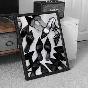 May include: Black and white framed print featuring a collection of women's shoes. The assortment includes various styles of heels, pumps, and sandals, with pointed toes and decorative elements. The shoes are arranged on a white background.