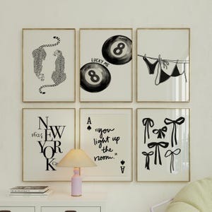 Puede incluir: Seis impresiones artísticas enmarcadas con marcos dorados. Las impresiones presentan ilustraciones en blanco y negro de guepardos, bolas de billar, lencería, el texto "NEW YORK", el texto "you light up the room" y lazos.