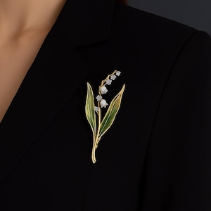 May include: A gold-coloured brooch shaped like a lily of the valley. It has green enamel leaves, white flower buds, and clear, sparkling crystals. The brooch is attached to a black blazer.