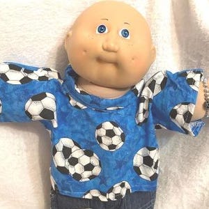 May include: A Cabbage Patch Kid doll wearing a blue shirt patterned with black and white soccer balls. The doll has blue eyes, a bald head, and a bracelet on its wrist. The shirt has a crew neck and short sleeves.