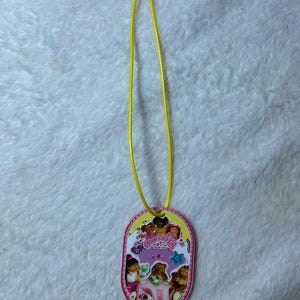 May include: An oval-shaped pendant featuring a Cabbage Patch Kids graphic with multiple characters, a pink table, and a purple background. The pendant has a bright yellow cord attached to the top, and the border is decorated with a pink and white stitch pattern.