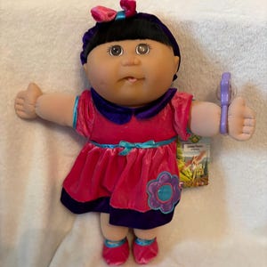 May include: A Cabbage Patch Kids doll with dark hair, a purple headband with a pink bow, and a bright pink velvet dress featuring a purple collar, a blue ribbon at the waist, and a large flower applique. The doll wears matching pink shoes.