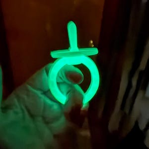 May include: A glow-in-the-dark pacifier featuring a bright neon green light emission. The object has a circular ring handle attached to a flat shield and a central nipple component, all illuminated with a vibrant, luminous green color in the dark.
