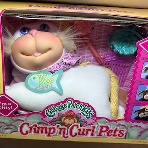 May include: A Cabbage Patch Kids Crimp 'n Curl Pets toy in a yellow box. The white plush kitty has blue eyes, a pink nose, and a pink patterned scarf. A teal fish-shaped tag is attached. Accessories include a pink comb and a teal hair accessory.