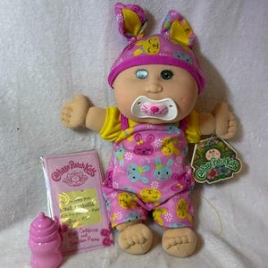 May include: A Cabbage Patch Kids doll wearing a pink bunny hat and overalls with bunny and flower patterns. The doll has a pacifier, and a pink bottle and birth certificate are included. The doll has a tag that says "Cabbage Patch Kids".