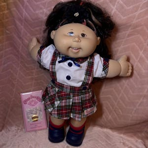 May include: A Cabbage Patch Kids doll with dark hair, a black flower headband, and heterochromia eyes. The doll wears a white short-sleeved shirt with a blue bow, a red and green plaid jumper, and navy blue shoes. A pink birth certificate card is included.