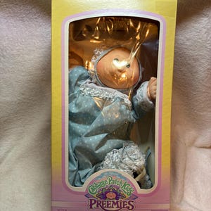 May include: A Cabbage Patch Kids Preemies doll in a yellow box with a clear plastic window. The doll has a round face, green eyes, and wears a light blue long-sleeved outfit with a white floral pattern, a matching bonnet, and white lace trim.