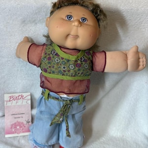 May include: A Cabbage Patch Kids doll with brown hair, blue eyes, and freckles. The doll wears a sheer maroon short-sleeve top with a green floral vest, light blue denim pants with a matching tie belt, and red shoes. A birth certificate is included.