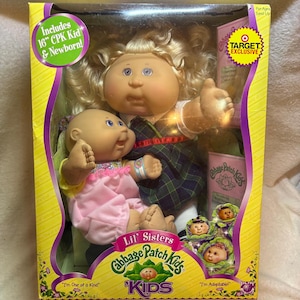 May include: A Cabbage Patch Kids Lil' Sisters boxed set featuring a 16-inch doll with blonde hair and a plaid dress, accompanied by a newborn doll in a pink outfit. The packaging includes Target Exclusive branding and Cabbage Patch Kids logos.