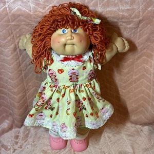 May include: A doll with curly auburn yarn hair, blue eyes, and freckles, wearing a pale yellow dress featuring a Strawberry Shortcake print, a white lace collar with a small red bow, and pink shoes. The doll has outstretched arms.