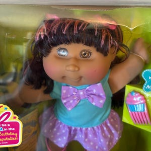 Puede incluir: Muñeca Cabbage Patch Kids con cabello castaño y mechas rosas, vestida con una camisa azul con un lazo morado, una falda morada con estrellas blancas y zapatos rosas. La muñeca tiene dos ojos de diferente color. La caja tiene texto "Be a R in the Birthday Keepsakes!"