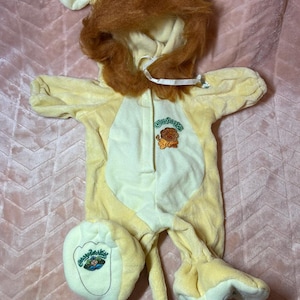 May include: A pale yellow plush lion costume for a doll, featuring a hood with a brown faux fur mane, long sleeves, and a tail. The chest displays an embroidered lion graphic with the text Cabbage Patch Kids, and one foot has a Cabbage Patch Kids logo.