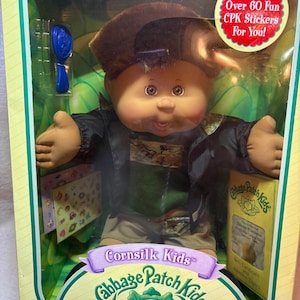 May include: A Cabbage Patch Kids Cornsilk Kids doll in original packaging. The doll has brown hair, a black jacket, a green shirt, and tan shorts. Included are a blue hairbrush, a sticker sheet, and adoption papers. The box features the text Includes Over 60 Fun CPK Stickers For You! and One of a Kind. And Just Mine!