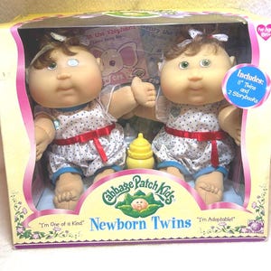 May include: A Cabbage Patch Kids Newborn Twins set in original packaging, featuring two 11-inch dolls with brown hair, green eyes, and white polka-dot outfits with red ribbons. Includes two storybooks and a yellow bottle inside the box.