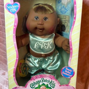 Könnte beinhalten: Eine Cabbage Patch Kids Newborn Puppe mit dunkler Haut, braunen Augen und blondem Haar, bekleidet mit einem hellblauen Satin-Lätzchen mit den Buchstaben CPK und passender Pumphose. Die Puppe ist in einer Schachtel mit der Aufschrift Newborn CPK verpackt.