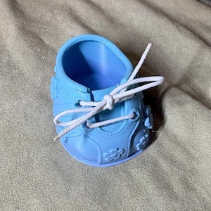 May include: A light blue shoe with a hollow interior, featuring a white lace tied in a bow across the top. The exterior surface of the shoe is decorated with small, raised flower shapes and has a structured, rounded toe and heel design.